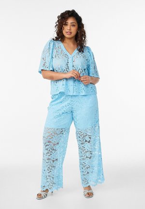 Kvinde iført lyseblå blonder kortærmet bluse og matchende bukser med vide ben, stående med hænderne blidt foldet, neutral baggrund.