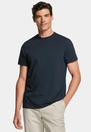 Homme portant un t-shirt ajusté à manches courtes bleu marine foncé et un pantalon beige clair, debout avec une main dans la poche devant un fond uni.