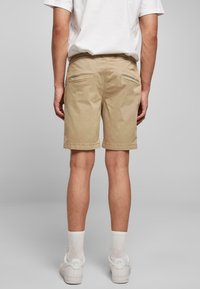 Short beige en mélange de coton texturé, présentant une coupe décontractée, des poches arrière et une ceinture. Portés avec des baskets blanches et des chaussettes.