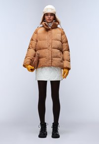 Brązowa kurtka puchowa z korduroy, biała pikowana mini spódnica, musztardowe rękawiczki, czarne sznurowane botki oraz puszysty biały kapelusz z nausznikami.