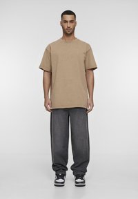 Camiseta marrón de gran tamaño con mangas cortas, combinada con jeans oscuros de corte holgado y zapatillas en blanco y negro. Diseño sencillo y casual.