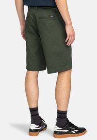 Grüne Shorts aus Baumwolle mit geradem Schnitt, zwei Gesäßtaschen und einem Logo-Patch. Kombiniert mit schwarzen Sneakers und gestreiften Socken.