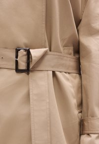 Μπεζ παλτό τύπου τραντς με ζώνη, που διαθέτει ορθογώνια σκούρα αγκράφα και ραμμένες λεπτομέρειες. Λείος υφής, κανονικό γιακά και πλαϊνή τσέπη.