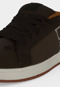 Baskets marron foncé avec des lacets noirs épais, une semelle intermédiaire blanche et une semelle extérieure en caoutchouc marron, montrant les détails avant et latéraux.