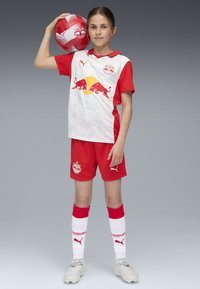 Röd och vit fotbollsklädsel med kortärmad tröja, shorts och knä-långa strumpor. Innehåller Red Bull-grafik och Puma-logotyper. Håller en röd boll.