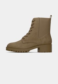 Bottine à lacets beige avec talon épais et languette, dotée d’un panneau latéral élastique et d’une semelle à crampons robustes, vue de profil sur un fond clair.