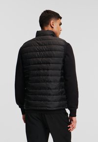Vesta puffer neagră cu un model matlasat și guler înalt, asociată cu un top negru cu mânecă lungă, văzută din spate.