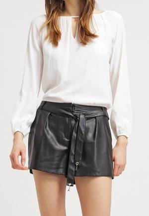 Frau trägt eine weiße Langarmbluse und hoch taillierte schwarze Leder-Shorts mit einem gebundenen Gürtel in der Taille.