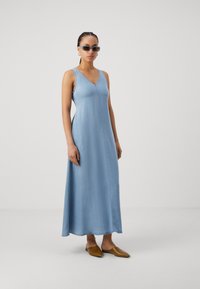 Rochie maxi din denim albastru deschis, cu decolteu în V și design fără mâneci, având o cusătură frontala și fante laterale. Purttă cu pantofi slip-on maro.