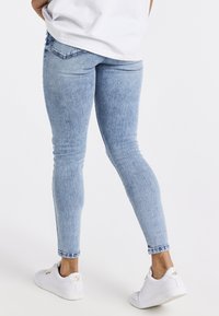 Persona con jeans ajustados de color azul claro y zapatillas blancas, de pie con las piernas ligeramente cruzadas frente a un fondo blanco liso.