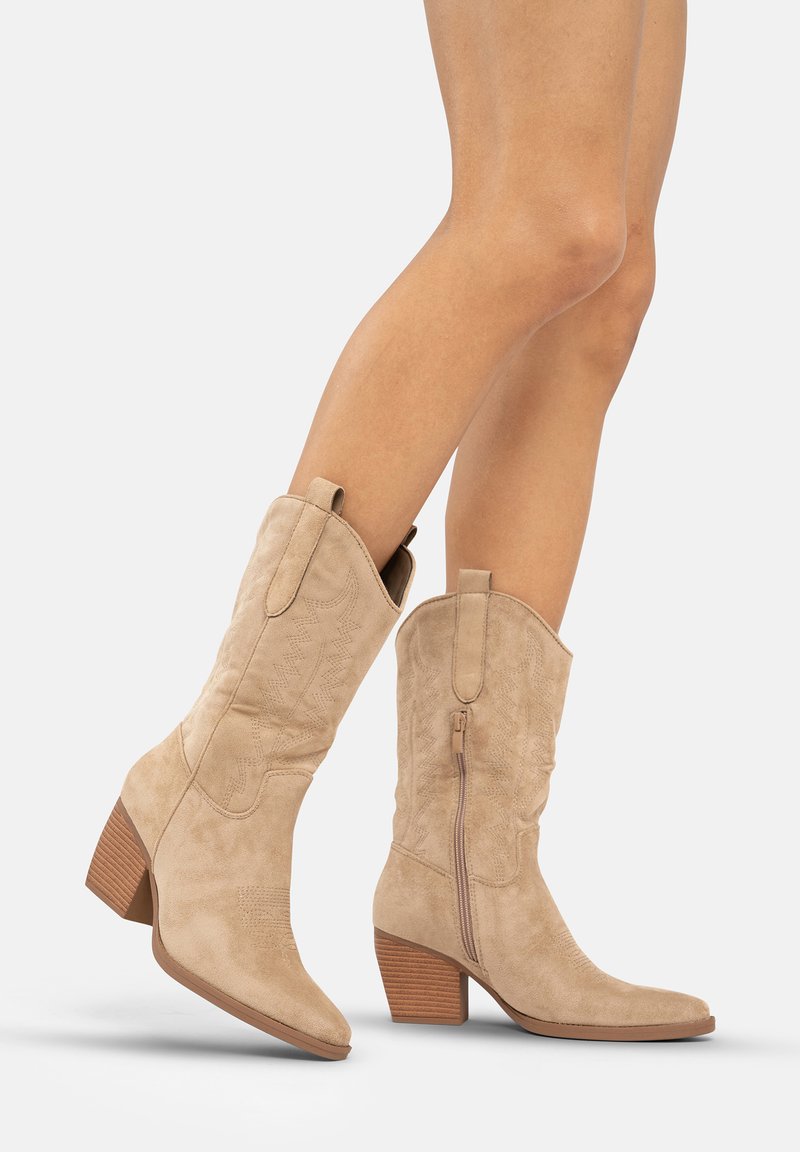 Botas de cowboy de ante beige con punta afilada, costuras decorativas y cremallera lateral. Presentan un tacón apilado y acentos texturizados.