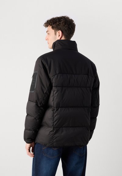Doudoune noire avec un col haut, présentant un design matelassé et un patch logo sur la manche gauche. Associée à un jean en denim avec une finition mate.