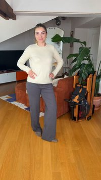 Femme debout, mains sur les hanches, dans un salon près d'un canapé marron, d'une grande plante verte et d'un rameur en bois sur un sol en parquet.