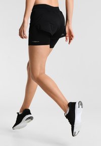 Pantalones cortos deportivos negros con tejido texturizado, cintura elástica y detalles reflectantes. Combinados con calzado negro con suela blanca.