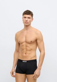 Fekete boxeralsó márkás elasztikus derékrésszel. Puha anyagból készült, testhezálló szabású és sima textúrájú, semleges háttér előtt egy modellen bemutatva.