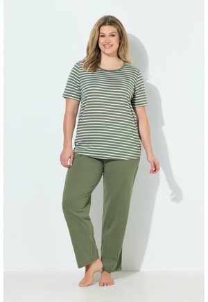 Smilende kvinde står barfodet, iført olivengrønne bukser og en kortærmet oliven- og hvidstribet skjorte mod en ensfarvet hvid baggrund.