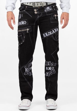 Mann trägt schwarze Jeans mit weiß besticktem Text und Designs, silberner Reißverschlusstasche, grauem Gürtel, weißen Sneakers und hellgrauem Sweatshirt.