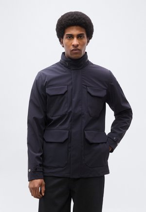 Moški z afro pričesko, oblečen v temno modro jakno s štirimi žepi z zavihki, stoji pred preprostim svetlim ozadjem, obrnjen naprej.