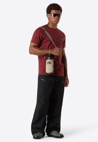 Mann mit dunklen Sonnenbrillen, kastanienfarbenem T-Shirt, schwarzen Hosen und camouflagefarbenen Schuhen, der eine beigefarbene Wasserflasche in einer Umhängetasche hält.