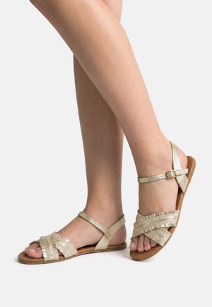 Gambe che indossano sandali piatti open toe con cinturini incrociati dorati e fibbia alla caviglia su sfondo bianco semplice.