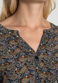 Schwarze Bluse aus Stoff mit einem Paisleymuster in Orange, Teal und Weiß, mit einem V-Ausschnitt und drei schwarzen Knöpfen auf der Vorderseite.