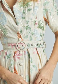 Abito ricamato in tessuto beige con motivi floreali in verde, rosa e blu. Con cintura decorata con una fibbia di legno circolare e bottoni frontali.