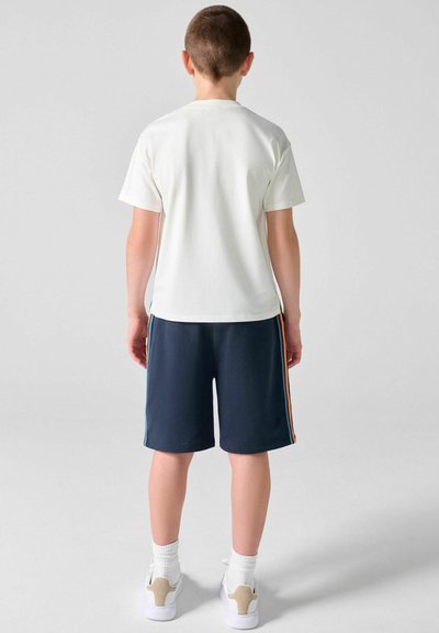 Garçon debout, de dos, portant un t-shirt blanc, un short bleu marine avec des bandes latérales, des chaussettes blanches et des baskets blanches sur un fond uni.