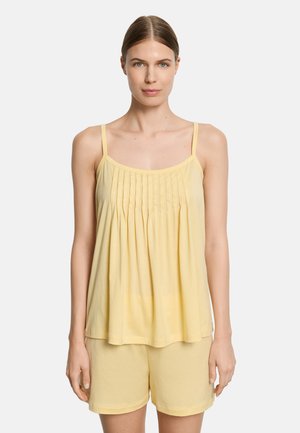 Vrouw die een lichtgele, geplooide, mouwloze top met dunne bandjes en bijpassende shorts draagt, staand tegen een effen achtergrond.