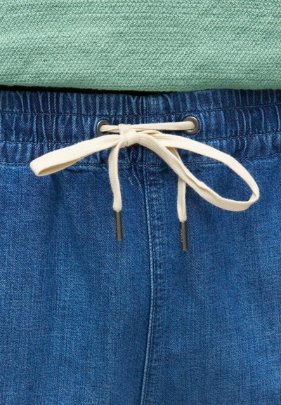 Κοντινό πλάνο μπλε τζιν παντελονιού με εκρού κορδόνι δεμένο σε φιόγκο πάνω σε φόντο από υφή πράσινου υφάσματος.