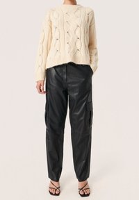 Jersey crema de punto grueso con un patrón texturizado, combinado con pantalones cargo de cuero negro con bolsillos laterales. Sandalias negras finas con tiras.