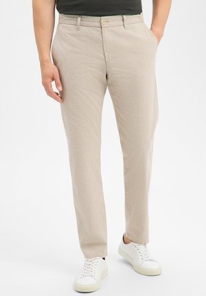 Man draagt beige geruite broek, zwart topje, witte sneakers, staat met één hand in de zak tegen een witte achtergrond.