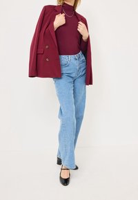 Burgundyröd blazer över en ribbad burgundyröd topp, kombinerad med ljusblå raka jeans och svarta klackar med ankelsnörning. Enkel bakgrund.