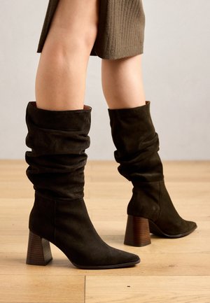 Botas tipo slouch de ante verde oscuro con punta afilada y tacón de madera apilada, que presentan una textura suave y un diseño de parte superior drapeada.