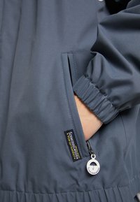 Veste légère bleu marine dotée de poignets élastiques, d'une poche zippée et d'une étiquette sur le côté avec un curseur de fermeture éclair rond en métal.