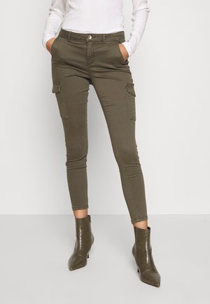 Donna che indossa pantaloni cargo aderenti verde oliva con tasche a patta e stivaletti alla caviglia texture coccodrillo verde oliva, in piedi con le mani in tasca.