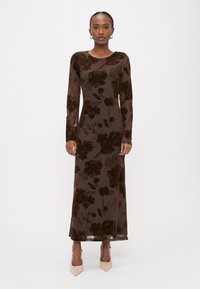 Μακρύ καφέ βελούδινο φόρεμα με λουλουδένιο σχέδιο, στρογγυλή λαιμόκοψη, μακριά μανίκια και ίσια σιλουέτα. Φορεμένο με μπεζ ψηλοτάκουνα παπούτσια μυτερά στη μύτη.