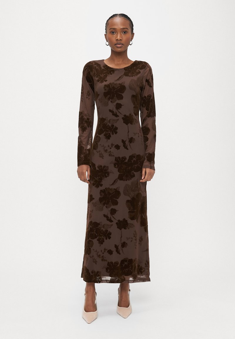 Μακρύ καφέ βελούδινο φόρεμα με λουλουδένιο σχέδιο, στρογγυλή λαιμόκοψη, μακριά μανίκια και ίσια σιλουέτα. Φορεμένο με μπεζ ψηλοτάκουνα παπούτσια μυτερά στη μύτη.
