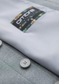 Chemise gris clair pliée avec une étiquette noire et colorée "Ombre", posée sur une surface en tissu gris texturé avec deux boutons ronds visibles.