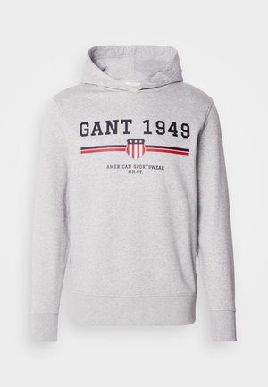 Szürke kapucnis pulóver, amelyen grafikus minta látható, vastag fekete betűkkel "GANT 1949" felirattal és egy amerikai pajzs logóval, piros és kék csíkokkal.