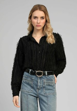 Femme blonde portant un chemisier noir texturé boutonné rentré dans un jean bleu taille haute avec de grandes poches devant et une ceinture noire.