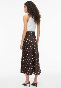 Brązowa spódnica midi w grochy wykonana z gładkiej tkaniny z rozkloszowanym dołem, zestawiona z białym topem na ramiączkach i brązowymi sandałami na obcasie.