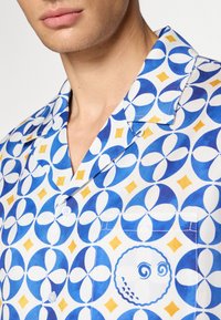 Polo majica s kratkimi rokavi iz bele tkanine, ki vsebuje modro in rumeno geometrijsko vzorčenje, z žepom na prsih, ki prikazuje krožno grafiko.