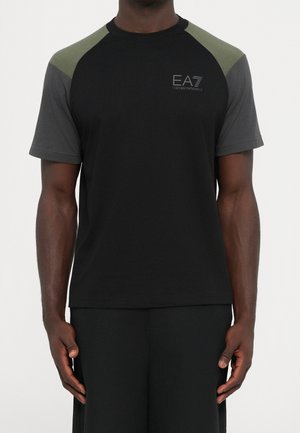 Uomo che indossa una maglietta nera a maniche corte con maniche raglan grigio scuro e verde oliva, con piccolo logo EA7 Emporio Armani sul petto.