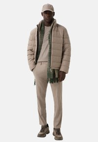 Man in een beige pufferjack, bijpassende broek en pet, een tan shirt, een groen geruite sjaal en beige-brown sneakers staat met zijn handen in de zakken.