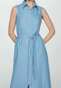 Sleeveless light blue denim dress with collar, button front, and waist tie, worn by a person with hands behind their back.