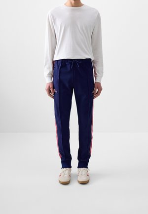 Personne portant une chemise blanche à manches longues, un pantalon de survêtement bleu marine avec des bandes rouges et des baskets blanches, debout devant un fond uni.