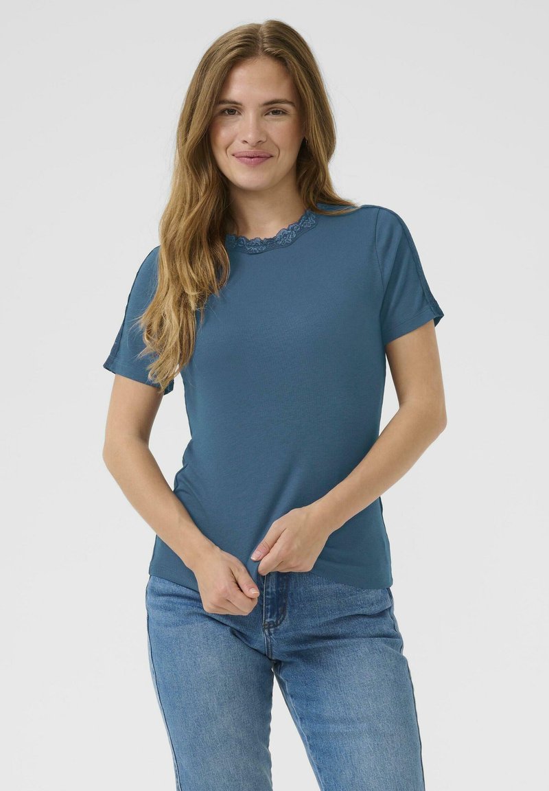Tealfarbener T-Shirt mit kurzen Ärmeln aus strukturiertem Stoff und rundem Halsausschnitt mit Borte. Getragen mit blauen Jeans, Hände, die das Shirt anpassen.