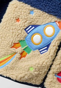 Fleece jopica s kremastim telesom in mornarsko modrimi poudarki, okrašena z vezenjem modre raketoplana in barvitimi zvezdami v rdeči, modri in zeleni barvi.