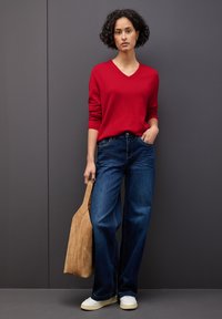 Maglione rosso a scollo a V, jeans a gamba larga blu, borsa in suede beige e sneakers bianche. Lo sfondo è grigio.