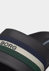 Μαύρο σανδάλι με λουρί από ύφασμα σε ρίγες μπλε ναυτικού, λευκού και πράσινου, με τη λέξη «BORG» σε μαύρα γράμματα.